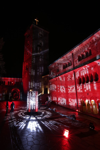 Das Bild zeigt den rot illuminierten zweiten Burghof, in dessen Mitte ein hoher, durchlöcherter Metallzylinder steht, der von innen heraus beleuchtet ist.