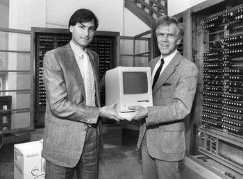 Das Bild zeigt Steve Jobs und einen anderen Mann, die einen der ersten Apple Macintoshs in den Händen halten.